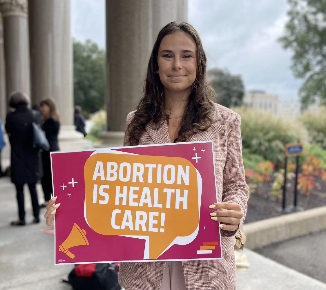 Jess, our policy counsel, is wearing a pink blazer holding a bright pink sign with an orange textbox that reads "ABORTION IS HEALTH CARE!" in white.