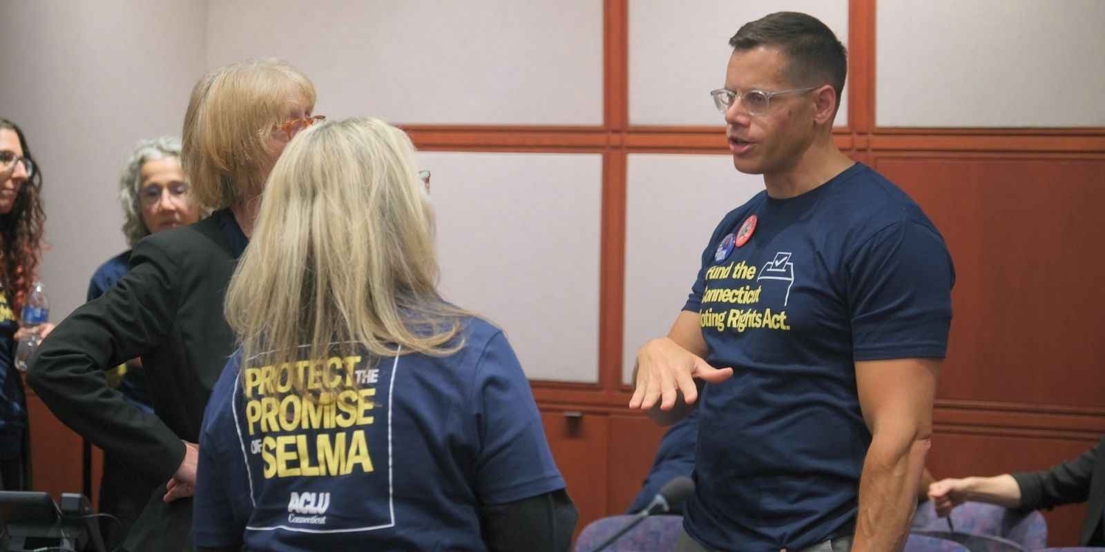 Gus, a white man with short blonde hair and glasses, is in a navy blue tshirt that reads in yellow: "Fund the Connecticut Voting Rights Act" on the front. He is talking with two white women who are blonde and wearing the same shirt for a lobby day.