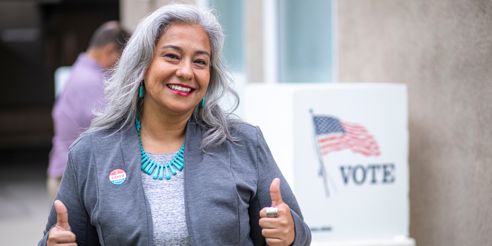 a person wearing a grey jacket smiling and giving a thumbs up. the person is wearing an "i voted" sticker.