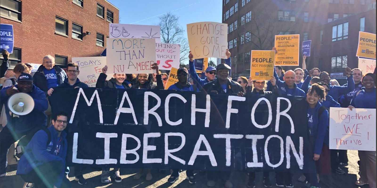 Our staff and Smart Justice Leaders are stood behind a navy blue banner that reads "MARCH FOR LIBERATION" with various posters.j