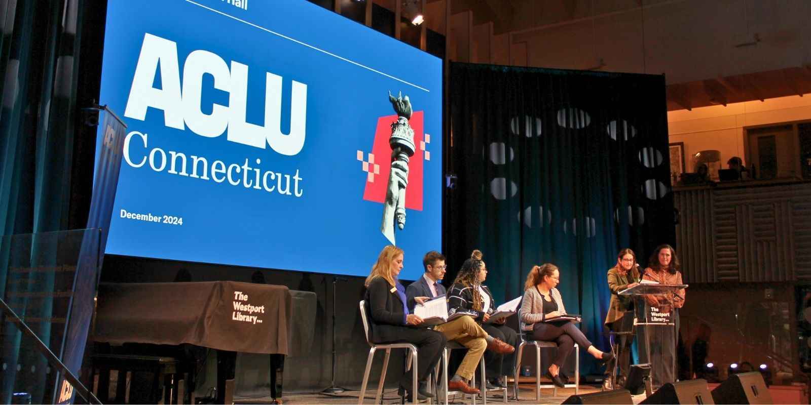 Staff members Amber, David, Chelsea-Infinity, and Elana sit on stage in front of a presentation with the ACLU of CT logo on a blue background before a full audience.