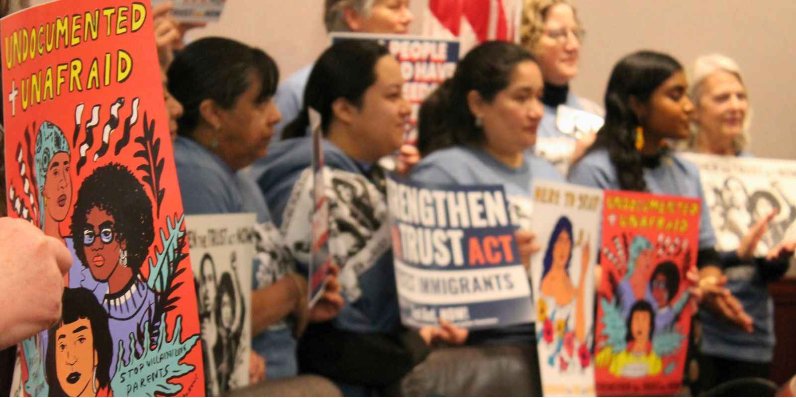 A group of women are standing in a hearing room at the Capitol, holding signs that says "Undocumented + Unafraid" and "Strengthen the Trust Act Now!"