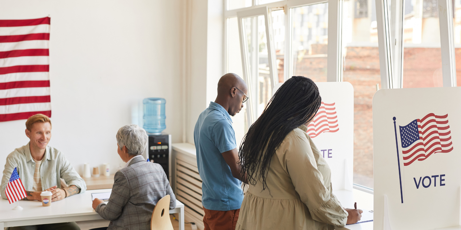 group of people at a polling location. Two are seated, chatting. Two are actively filling out ballots.