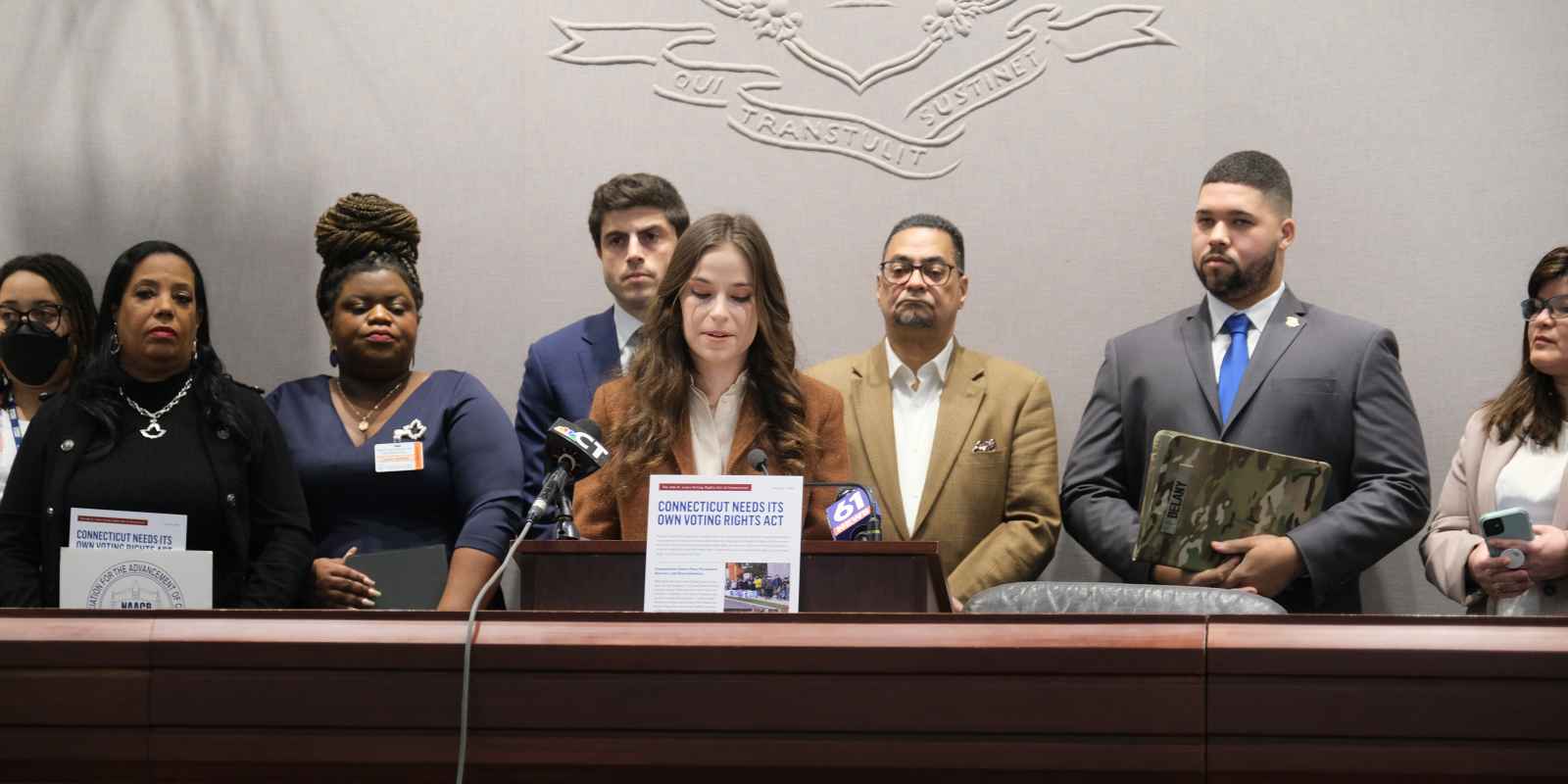 Jess Zaccagnino, a woman with long brown hair, stands behind a podium. In front of her is a sign that says "CT voting rights act." Behind her is a group of people, watching as she speaks