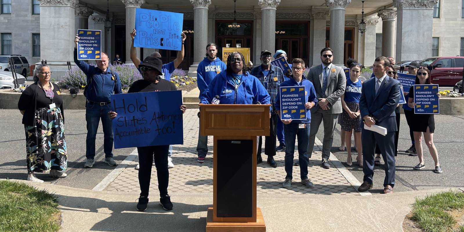Smart Justice leaders are gathered in front of the Capitol to speak about prosecutorial accountability and one leader, in the center, stands at the podium as they speak.