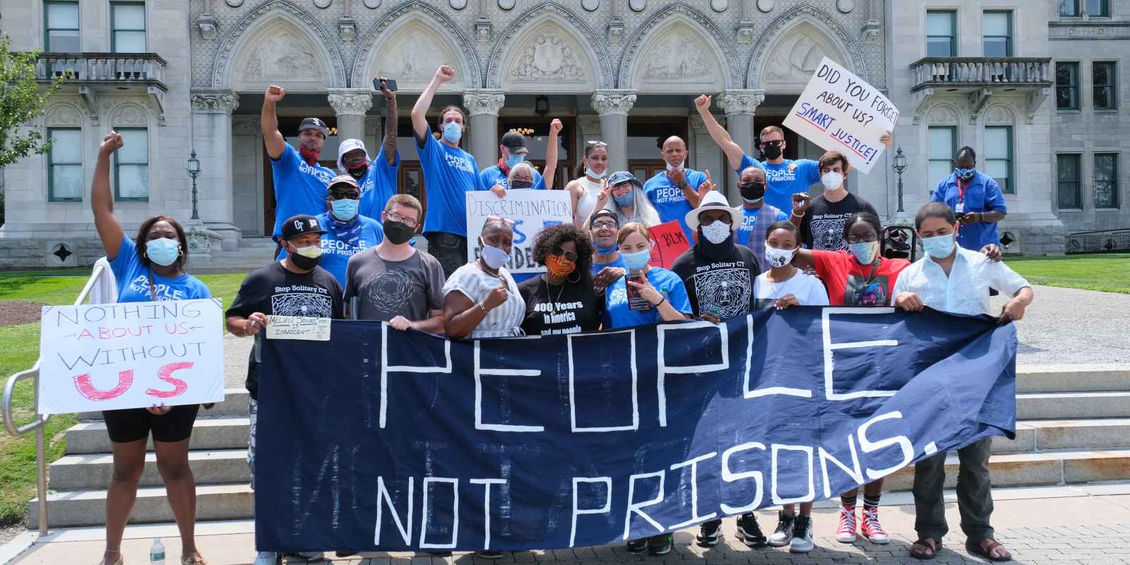 A group of Smart Justice leaders, advocates, and legislators, all wearing masks, stand behind the ACLU-CT's blue "People Not Prisons" banner outside of the Connecticut state capitol building.