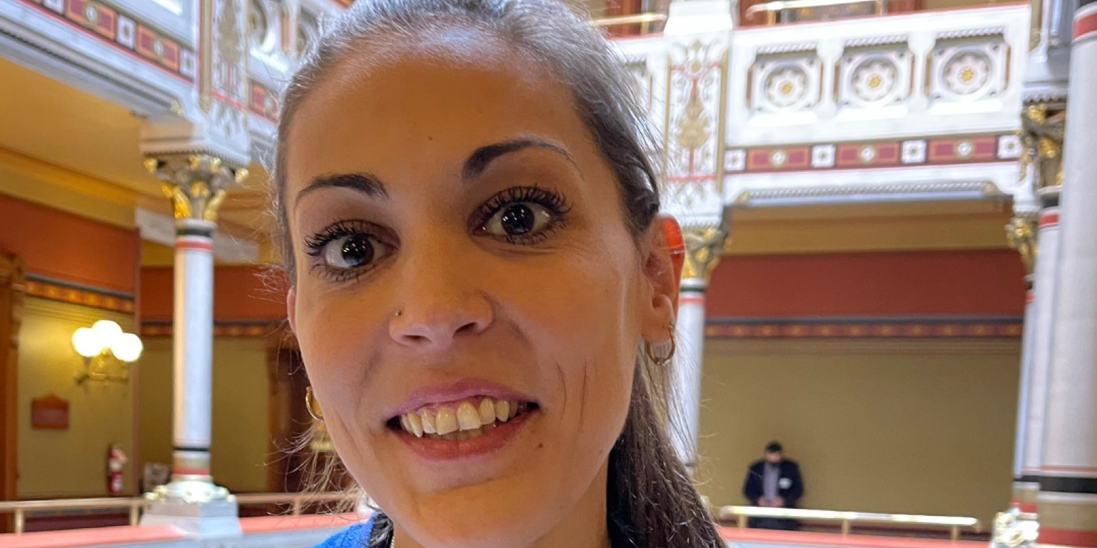 Image description: Alex Brown, ACLU-CT Smart Justice leader, stands, smiling directly at the camera. She is wearing a blue People Not Prisons t-shirt and black long-sleeved shirt under it. Her long brown hair is in a ponytail.