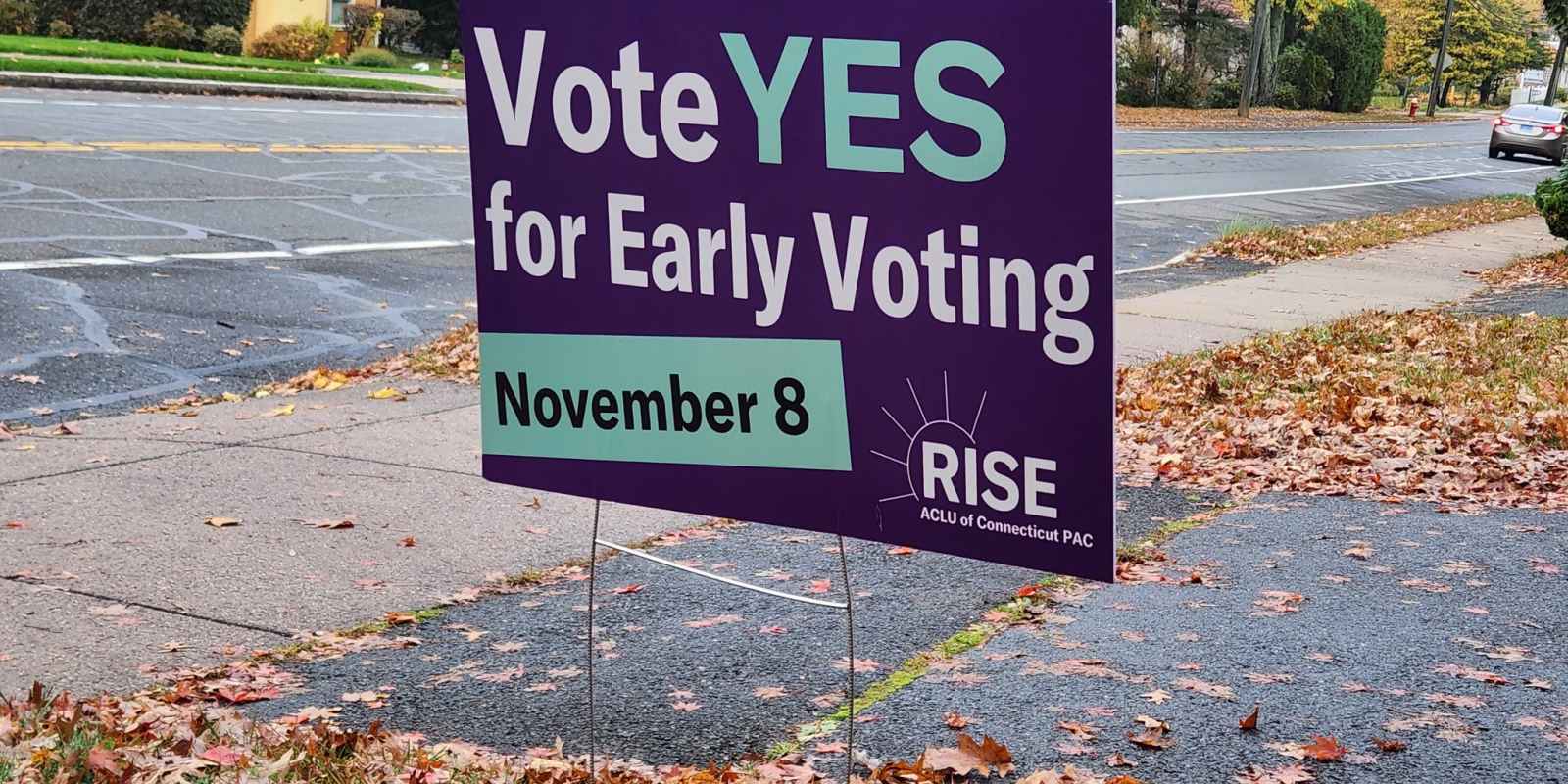 A purple yard sign, with green and white text, says, "vote YES for early voting November 8." the ACLU-CT Rise PAC logo is at the bottom. behind are a street and sidewalk, with autumn leaves scattered around