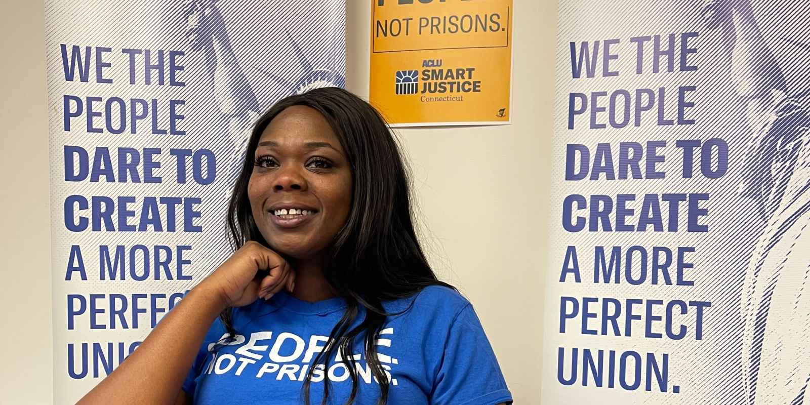 ACLUCT Smart Justice leader Shelby Henderson sits behind a desk, smiling at the camera, with one hand on her chin. She is behind a desk, laptop and wearing a blue people not prisons shirt, with ACLU of CT banners and a people not prisons poster behind her