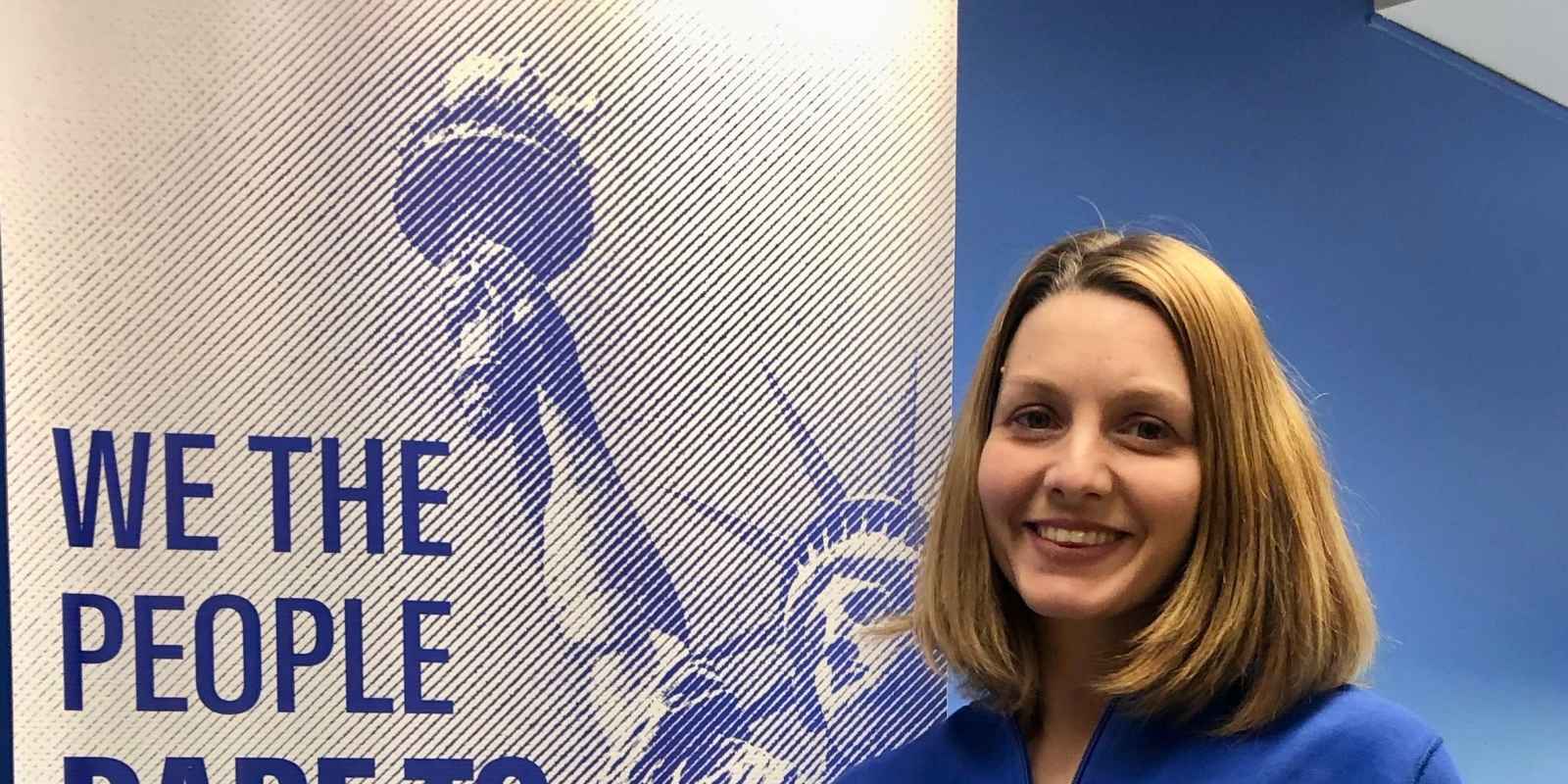 ACLUCT smart justice leader Tracie Bernardi stands facing the camera and smiling. She is wearing a blue Smart Justice zip-up and a yellow People Not Prisons pin. To her left is an ACLUCT banner that says we the people dare to create a more perfect union