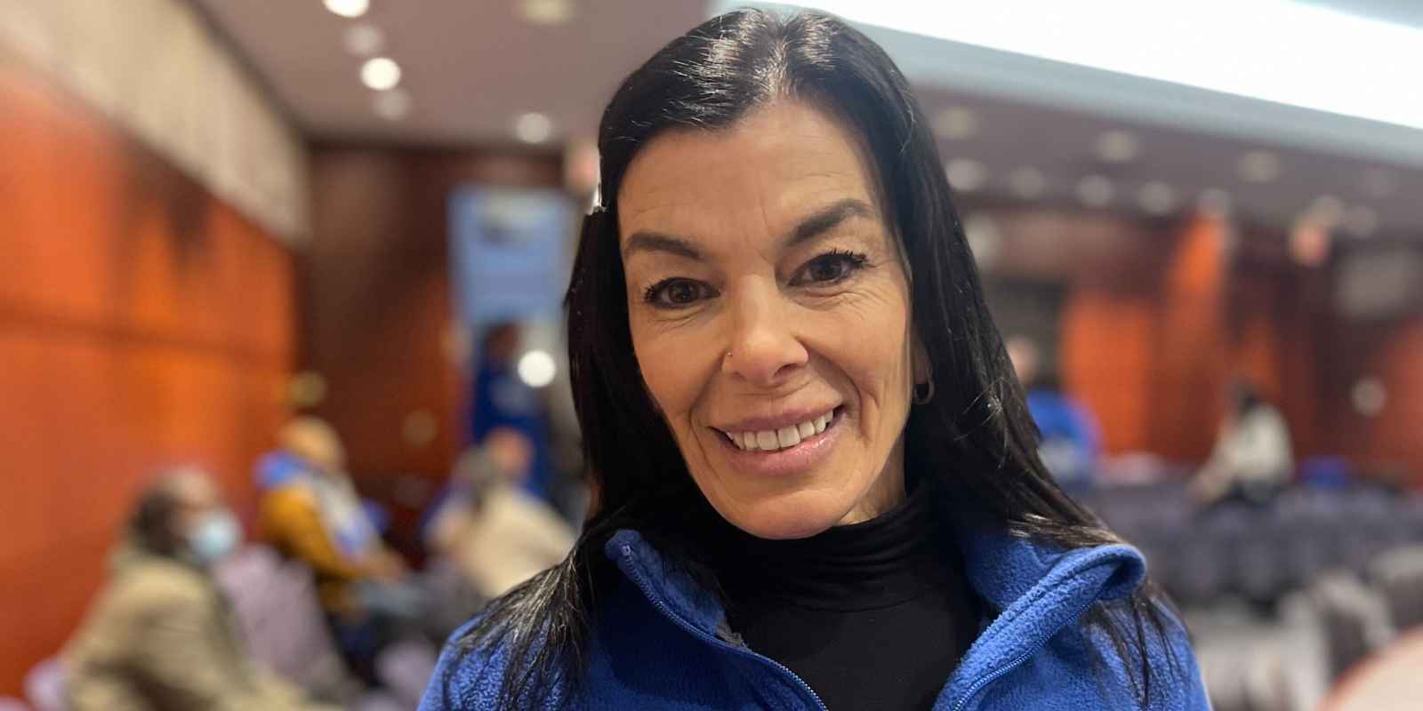 ACLU-CT smart justice leader Lori LeDonne stands, smiling at the camera and wearing a blue zip up sweatshirt. Behind her is a legislative hearing room