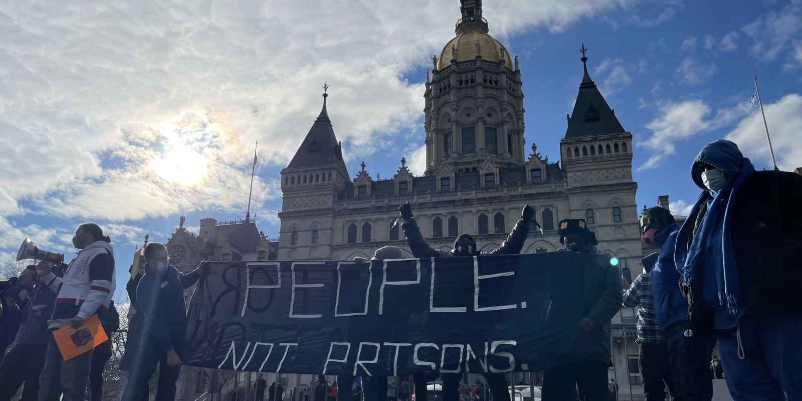 Smart Justice leaders stand in front of the CT capitol. The sun is breaking through a cloud. One person stands with arms upstretched above their head, behind the people not prisons banner