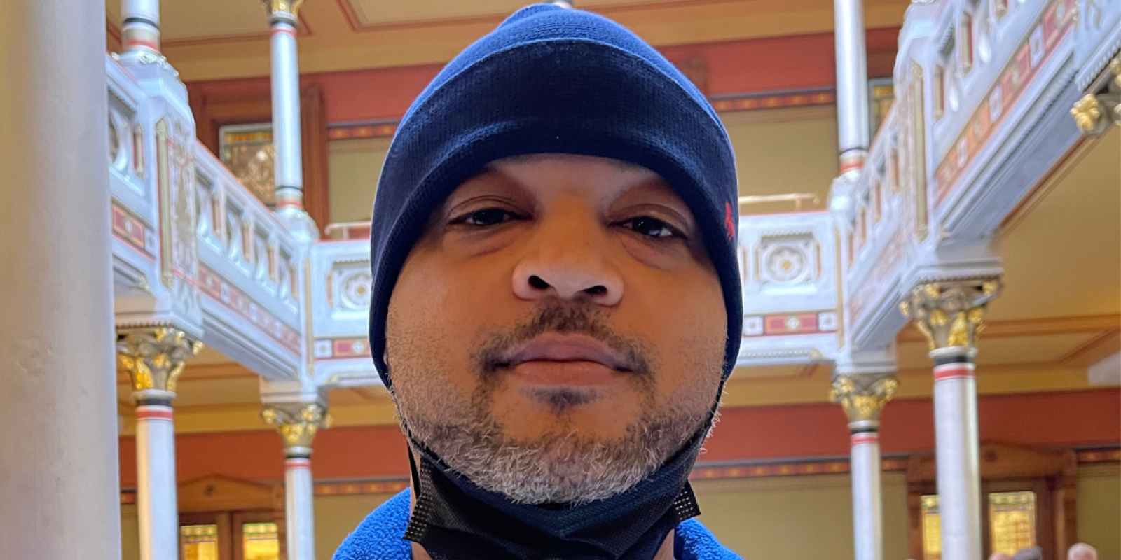 Image description: Tyran Sampson, ACLU-CT Smart Justice leader, stands, facing the camera. He is wearing a blue ACLU of Connecticut Smart Justice zip-up sweatshirt, a navy blue hat, and has a mask pulled down. He looks serious.