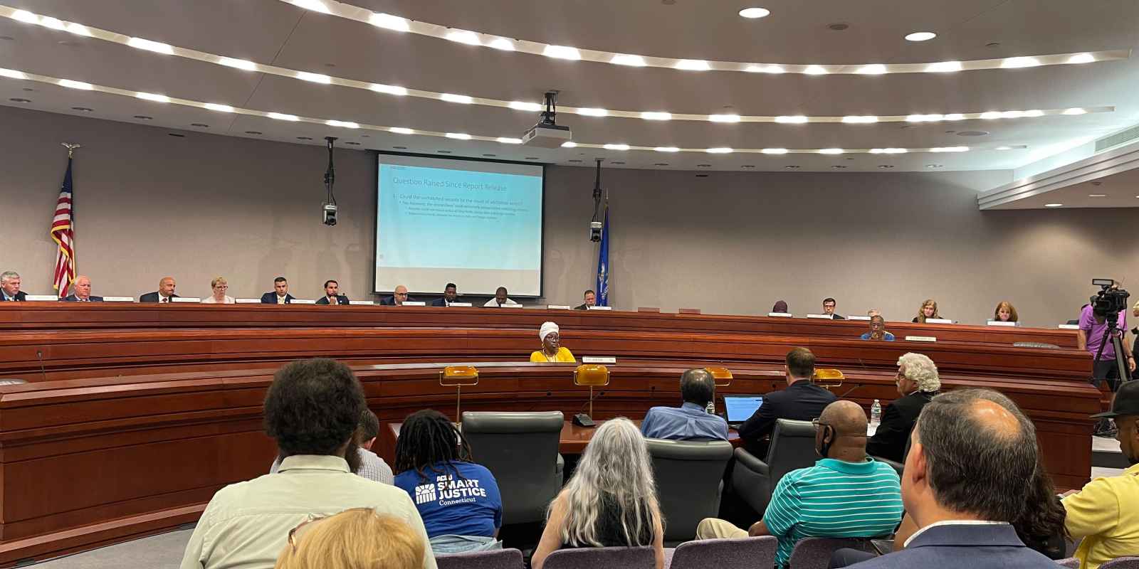 This photo captures a legislative forum about the fake traffic ticket scandal from the back of the room. Several people's backs are towards the camera with legislators in the background of the photo.