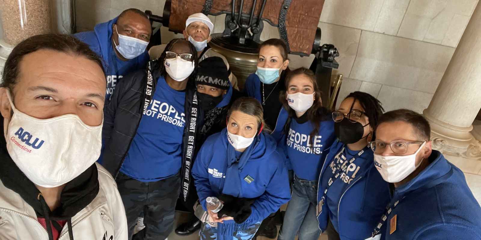 ACLU-CT Smart Justice leaders crouch in front of the bell at the CT State Capitol. They are wearing masks and blue people not prisons shirts, and smiling at the camera. 