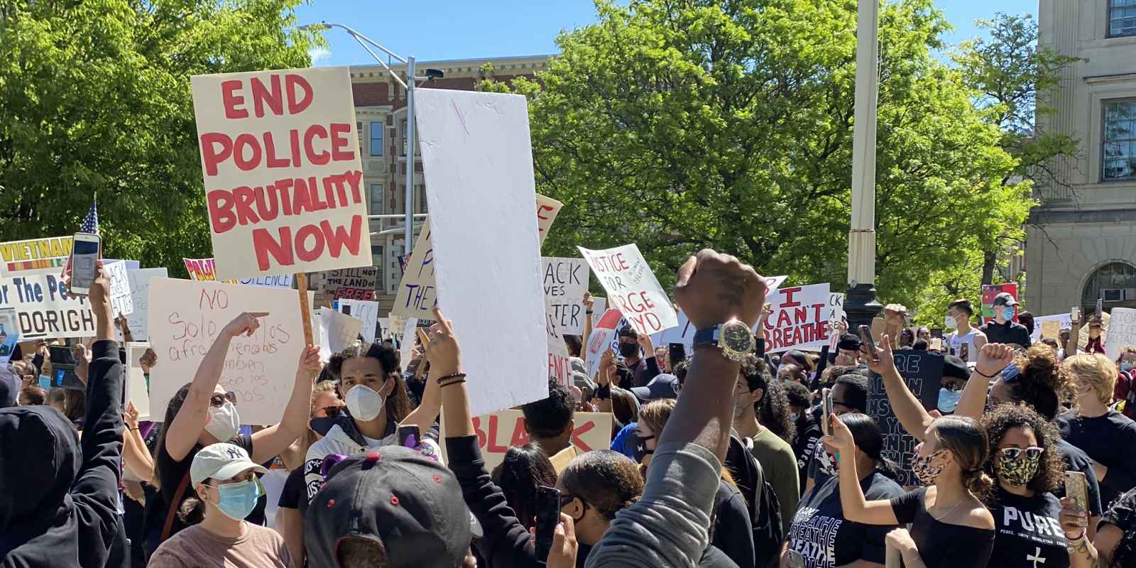 A crowd of protesters walk past the camera. The focus is on a sign that says "End Police Brutality Now."