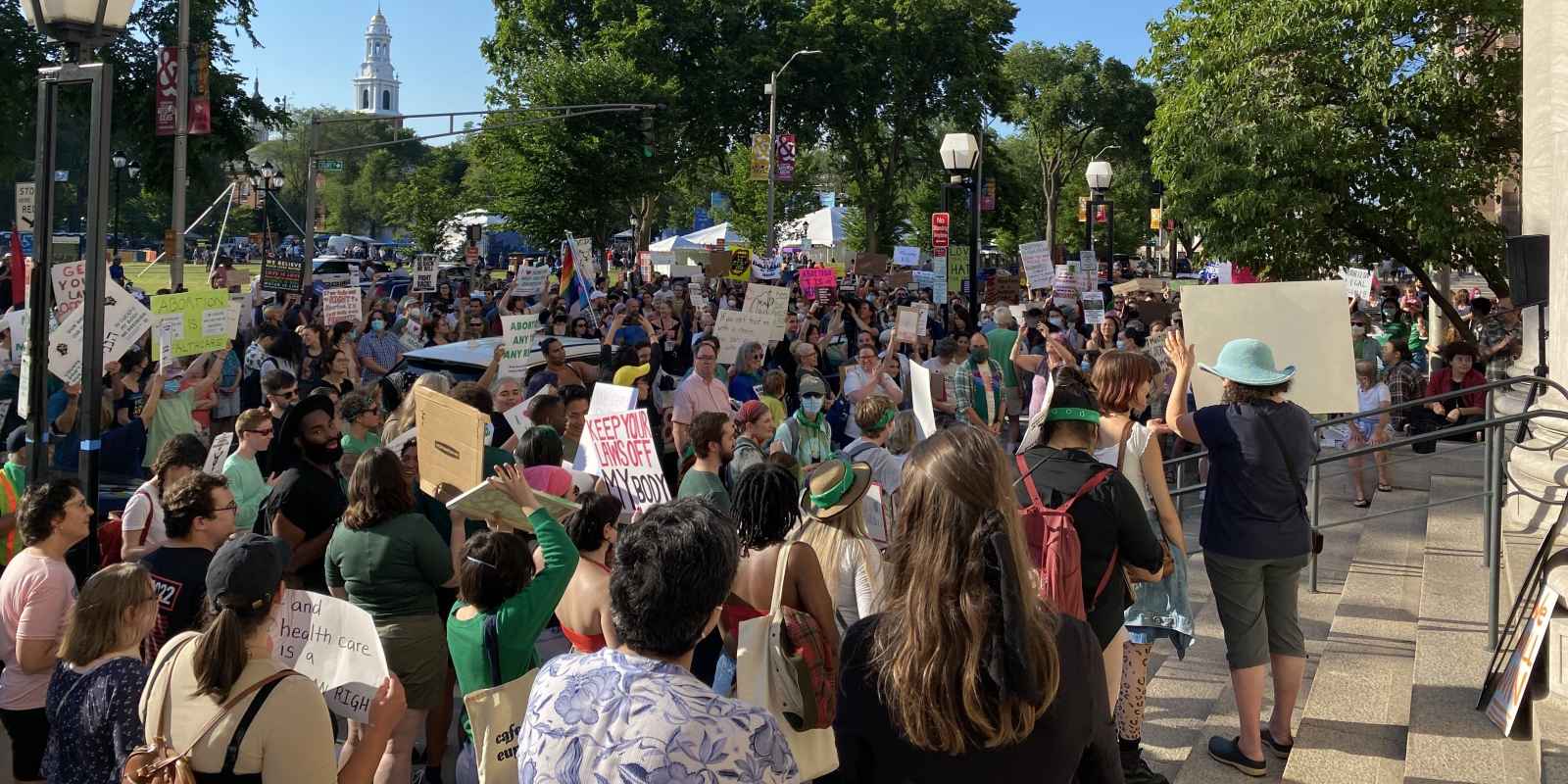 Protesters gather in New Haven after the US Supreme Court gutted Roe v Wade in the Dobbs decision. The sky is blue and clear, people are holding signs and trees are visible in the background