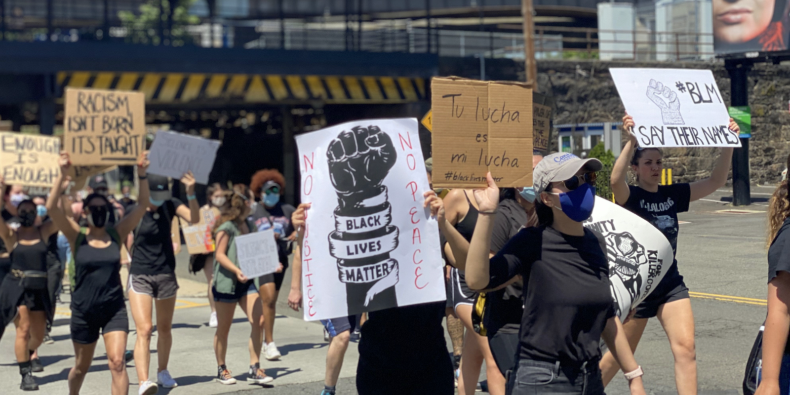 A crowd of protesters walk past the camera. The focus is on a sign that says," Black Lives Matter" around a fist.