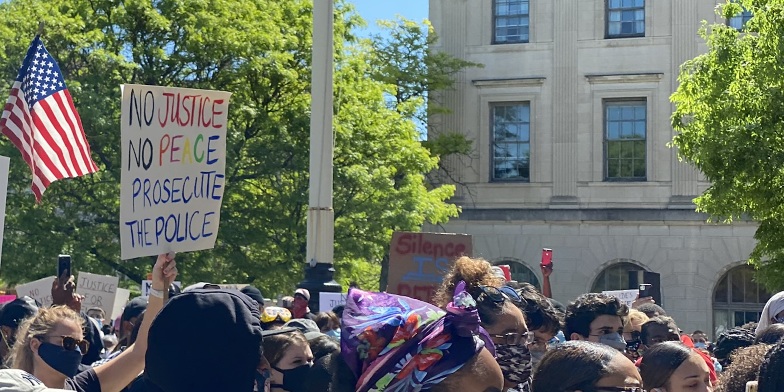 A crowd of protesters walk past the camera. The focus is on a sign that says "No Justice, No Peace, Prosecute the Police."