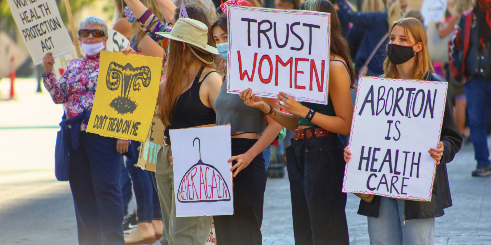 A group of women are the focus of the camera as they stand with signs that say, "Abortion is Healthcare" and "Trust Women."