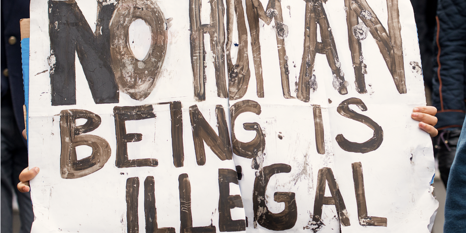 In front of a protest, a person is holding a big white sign that says "No human being is illegal."