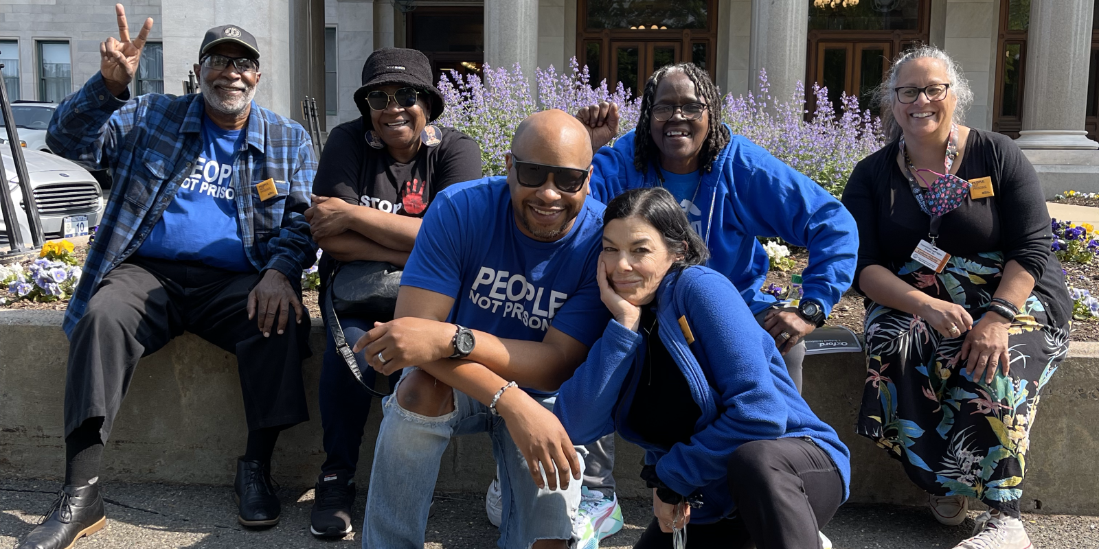 Six ACLU-CT Smart Justice leaders and friends sit and crouch in front of the CT Capitol building. They are smiling, wearing blue "people not prisons" shirts, and the sun is shining.