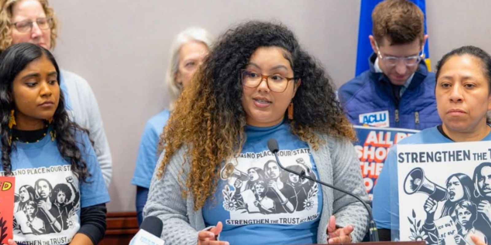 Chelsea-Infinity, our policy director, stands at the mic in a hearing room at the Capitol surrounded by Trust Act Now Coalition members holding signs that say "Undocumented + Unafraid" and "Strengthen the Trust Act Now."