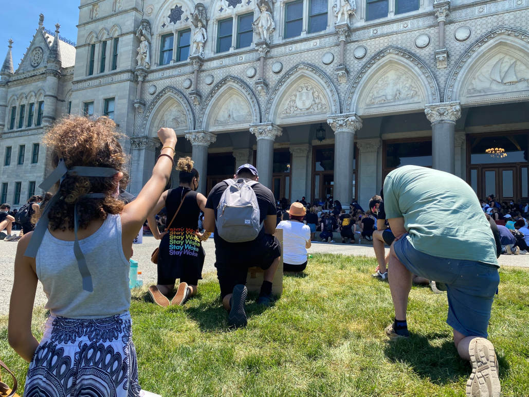 A crowd of people, spaced apart for physical distancing, take a knee in front of the Connecticut capitol building. A person in the foreground, hair up in a ponytail, holds her fist up.