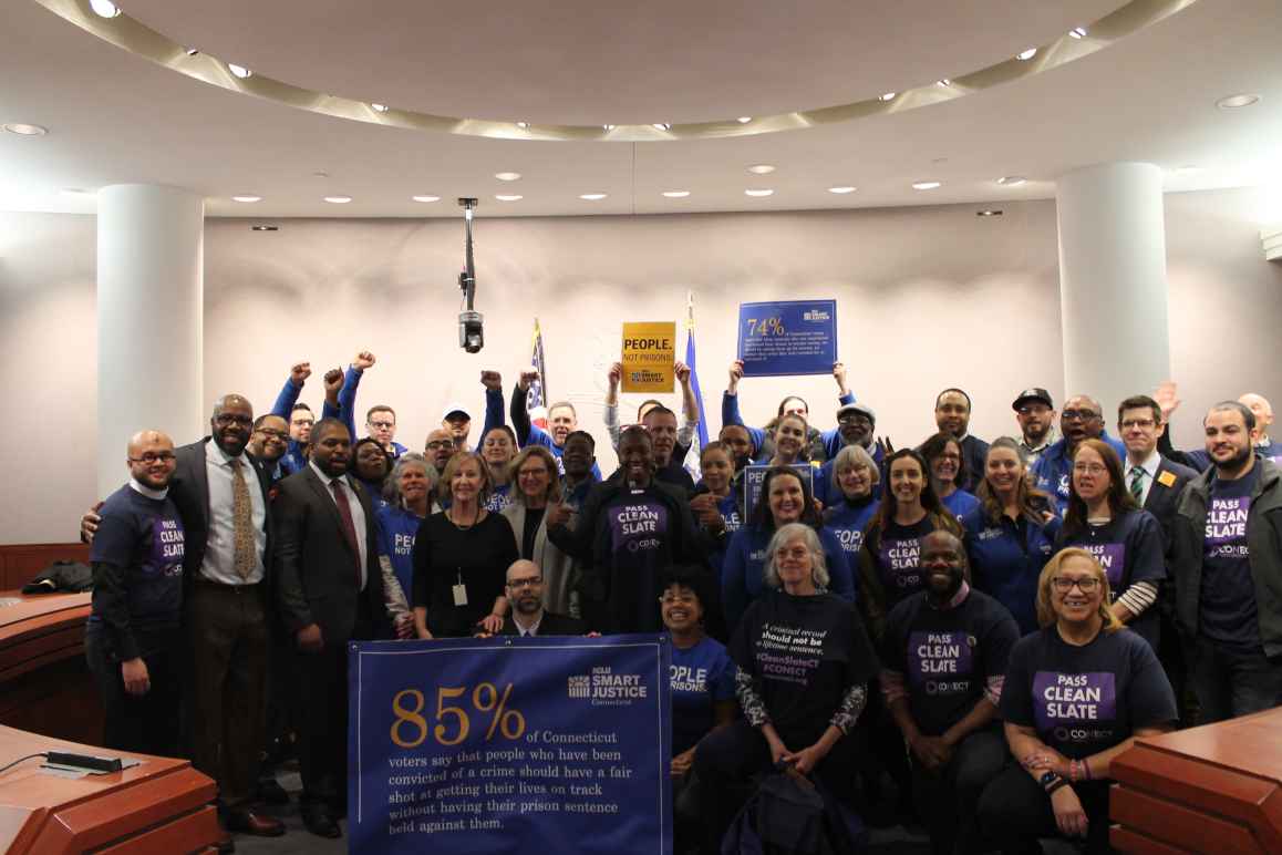 ACLU of Connecticut Smart Justice, CONECT, GHIAA, and Connecticut state legislators stand and sit in a group to support Clean Slate, behind a blue banner with a statistic about voters' support for Clean Slate ideas.