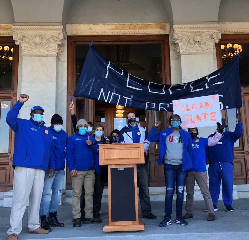 ACLU-CT Smart Justice leaders stand with "people not prisons" banner and "Clean Slate" sign outside of the CT State Capitol. They are behind a podium, and wearing blue "people not prisons" shirts. Many have fists up in power salutes.