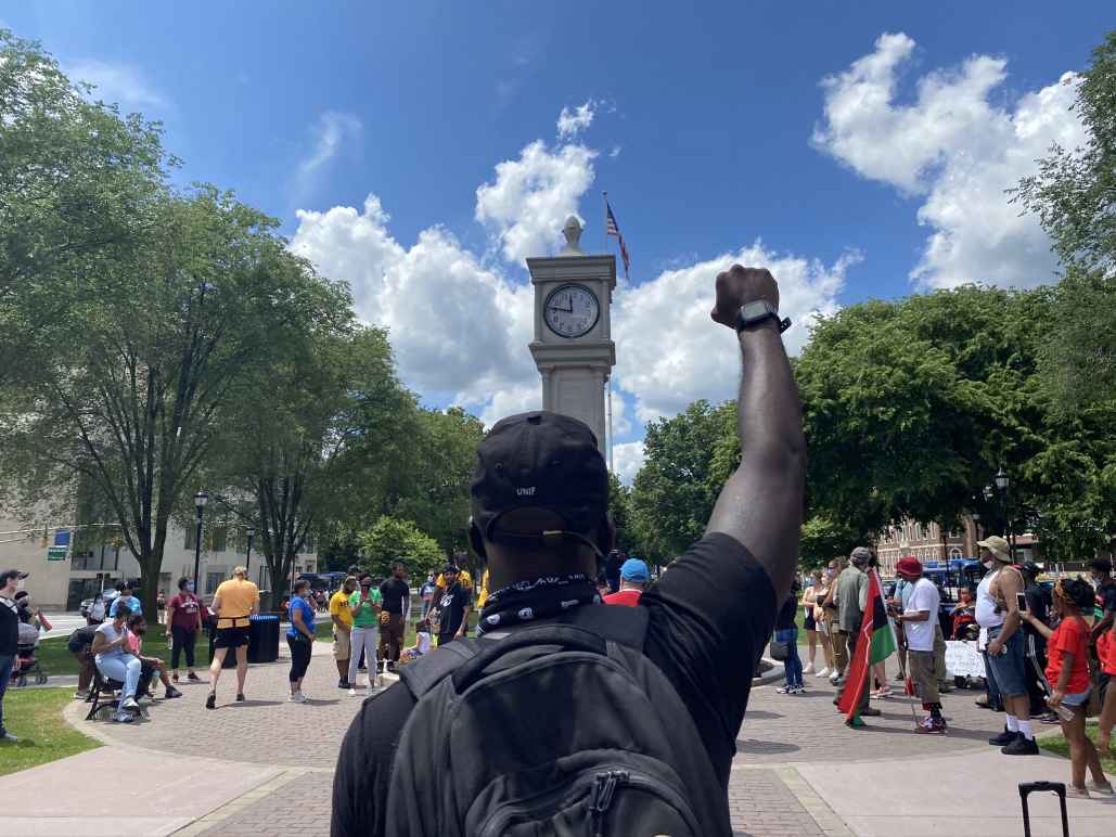 A crowd of people is gathered around the clock tower in Waterbury, Connecticut. The sky is blue and it is sunny. A Black man is in the foreground, back to the camera, with fist raised. He is wearing a backpack. 