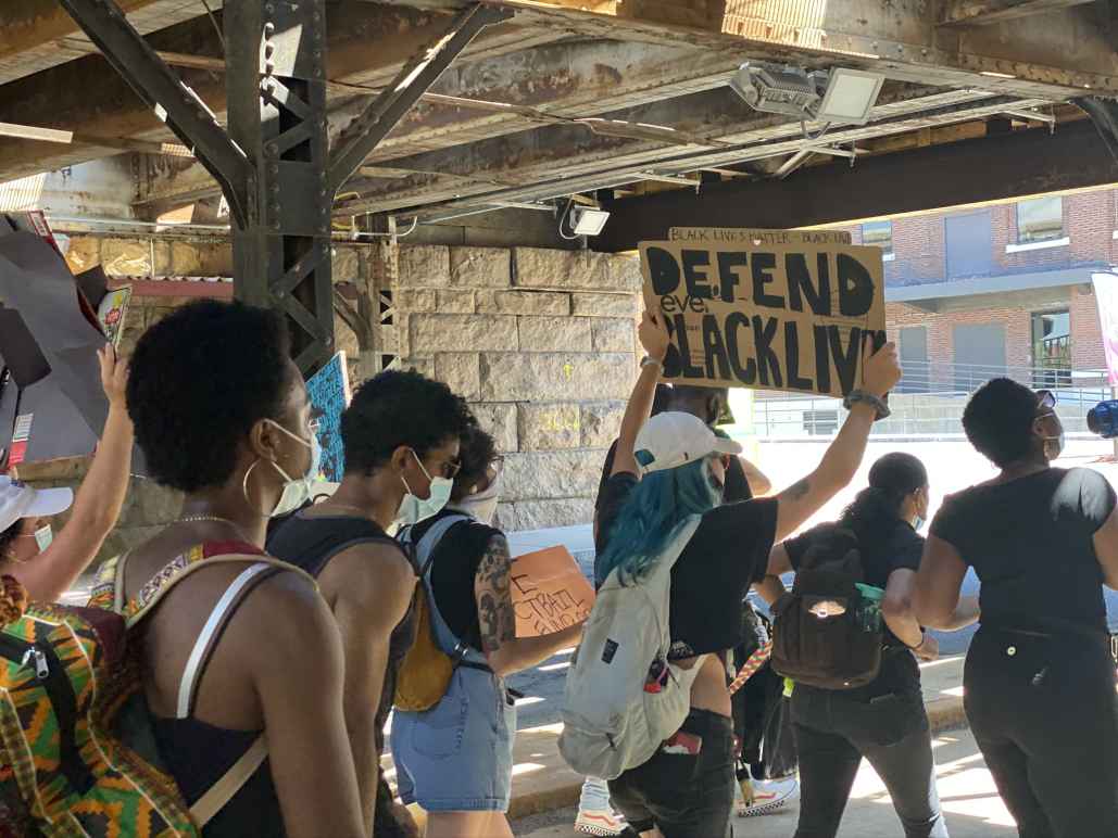 A group of protesters walks in Hartford. One holds a cardboard sign that says, in black letters, "defend Black lives"