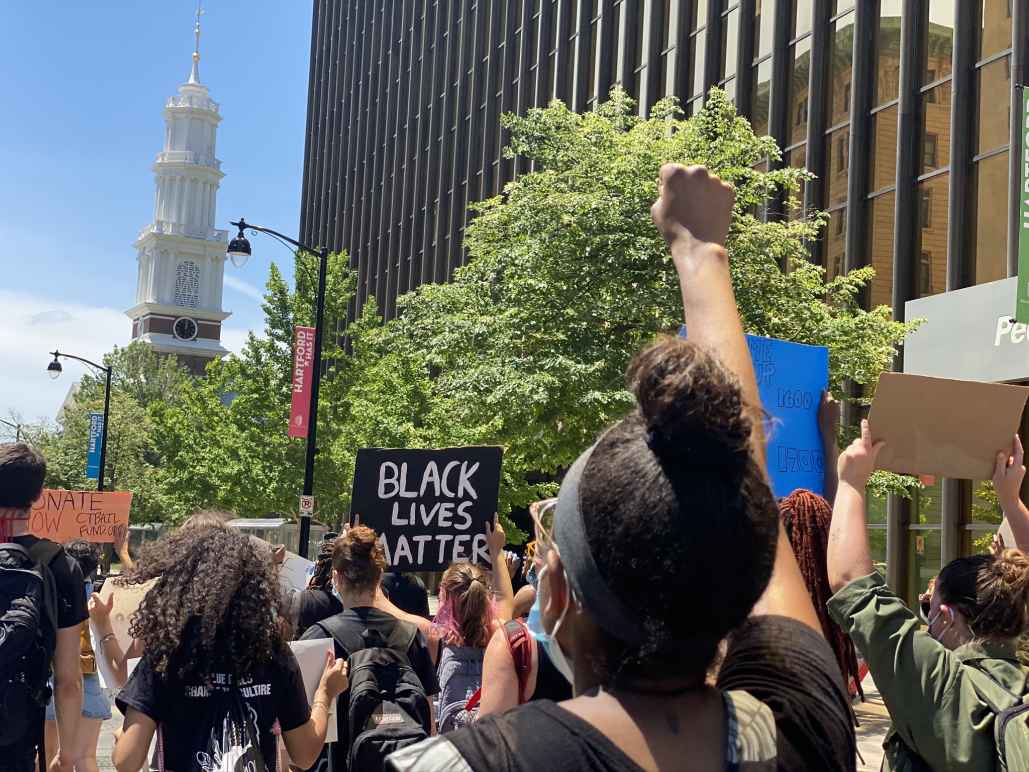 Protesters march in Hartford to call for the state to value Black lives. In the foreground, a person has their fist up. In the background, a Black Lives Matter sign