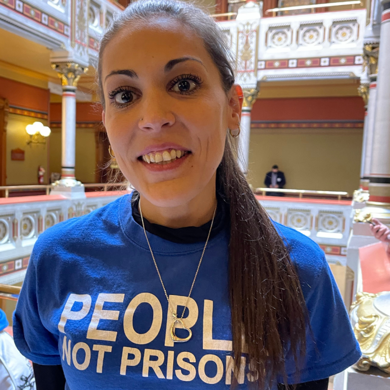 Image description: Alex Brown, ACLU-CT Smart Justice leader, stands, smiling directly at the camera. She is wearing a blue People Not Prisons t-shirt and black long-sleeved shirt under it. Her long brown hair is in a ponytail.