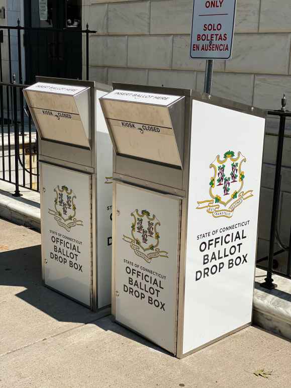 Two official Connecticut ballot drop boxes sit outside of the Waterbury city hall in 2020. The boxes are large, rectangular, and look like library book drops, with the state of CT seal visible, and "official ballot drop box," on their fronts and sides 