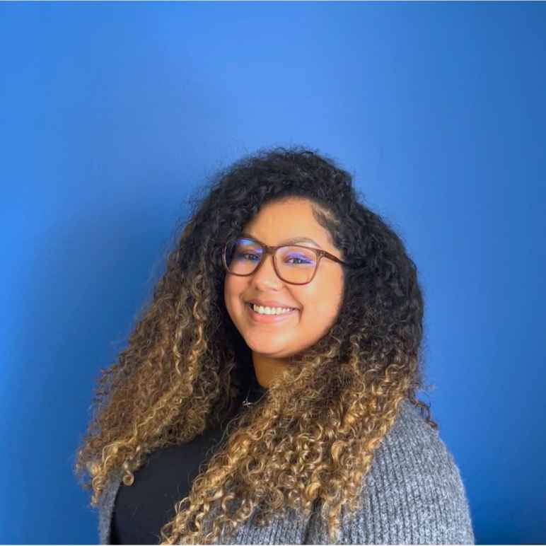 An Afro-Bori woman with curly hair, her roots are dark and her ends are bleached, in a grey cardigan smiles at the camera in front of a blue wall.