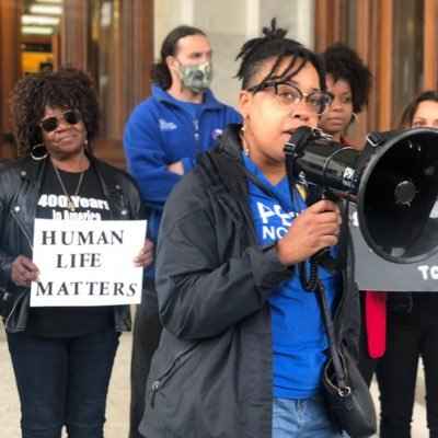 Claudine Fox ACLUCT public policy and advocacy director delivers a speech through a bullhorn. She wears a blue people not prisons hoodie and black coat. behind her, Barbara Fair holds a sign that says human life matters.