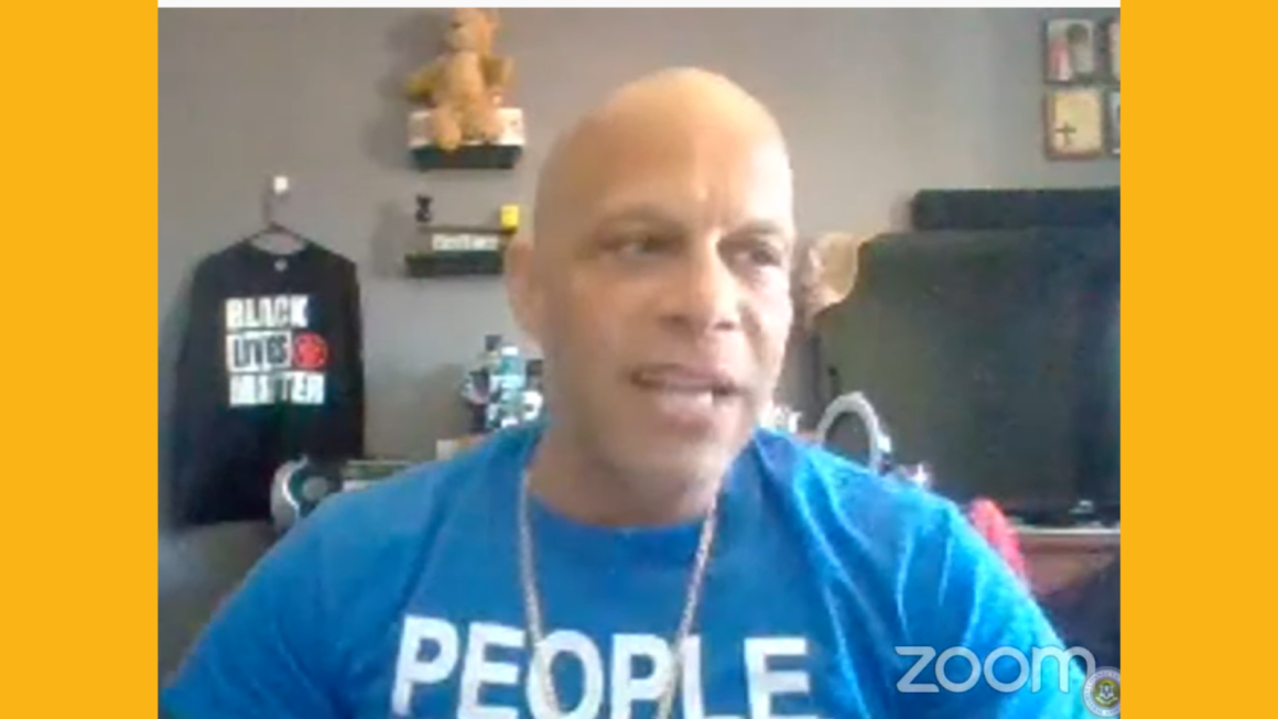 ACLU-CT Smart Justice leader Brian Sullivan testifies before the judiciary committee March 22, 2021. He is seated + wearing a blue people not prisons shirt, looking slightly to the right. Behind him is a Black Lives Matter shirt on a wall.