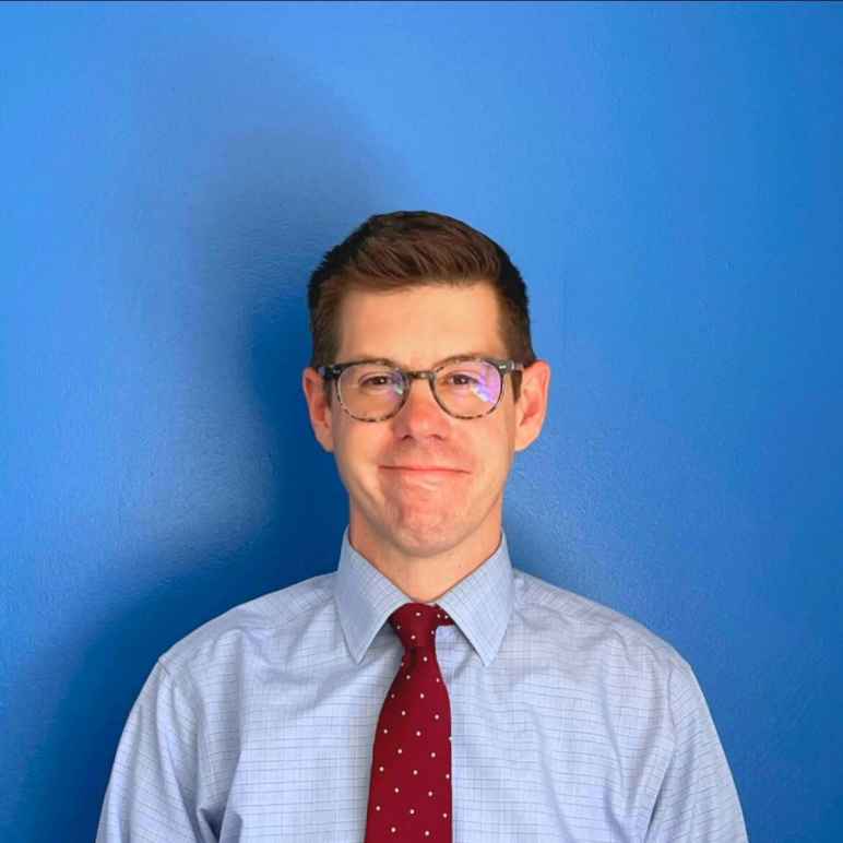 David McGuire, a white man with short brown hair wearing a light blue dress shirt with a red tie and brown glasses, looking at the camera with a smile.