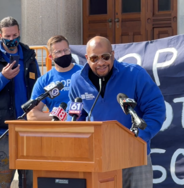Smart Justice leader Donald Rivers, in a blue zip up Smart Justice shirt and sunglasses, speaks at a podium during an April 21, 2021 press conference.