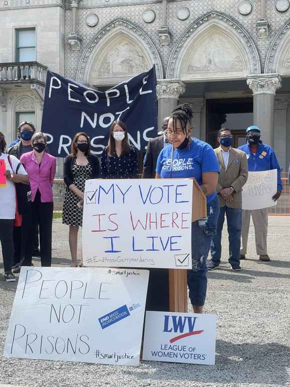 Claudine Fox, ACLU of Connecticut public policy and advocacy director, speaks at a press conference re ending prison gerrymandering. She's at a podium. In front of her is a sign: "my vote is where I live." Behind her, a group of legislators, masked.