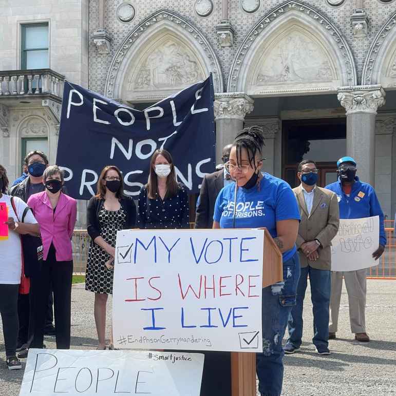 ACLU-CT public policy and advocacy director Claudine Fox, in a blue "people not prisons" smart justice shirt, speaks behind a podium in front of the CT capitol building. The podium sign says, my vote is where I live.