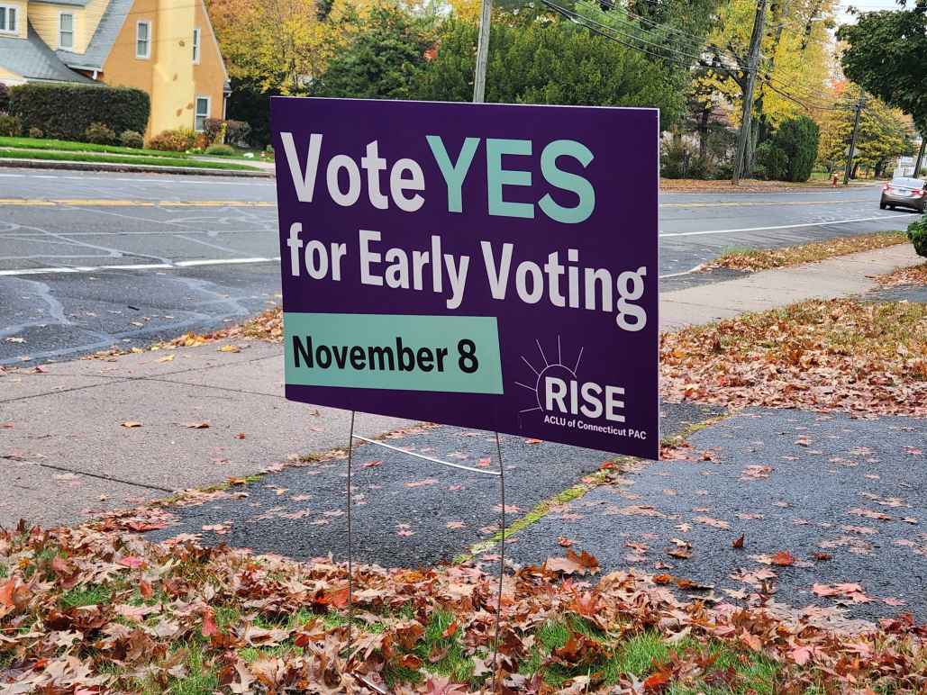 A purple yard sign, with green and white text, says, "vote YES for early voting November 8." the ACLU-CT Rise PAC logo is at the bottom. behind are a street and sidewalk, with autumn leaves scattered around