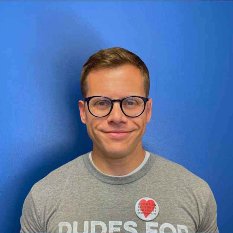 Gus Marks-Hamilton, a white man with black round glasses, smiles at the camera in front a blue wall with a gray shirt that says "Dudes for Abortions."