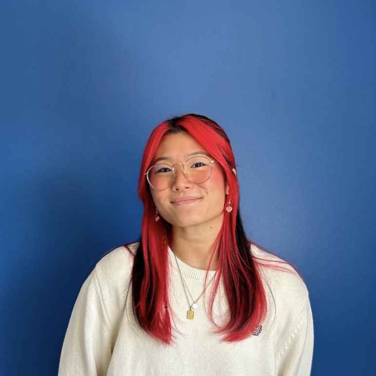 A Korean woman with long black hair, with red dyed hair in the front of and under her black hair is smiling at the camera. She is wearing gold-rim glasses and a white sweater on a blue background.