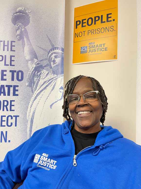 ACLUCT smart justice leader Terri Ricks sits, smiling and looking straight at the camera. She is wearing a blue ACLU of Connecticut Smart justice hoodie and glasses. Behind her is a yellow people not prisons poster.