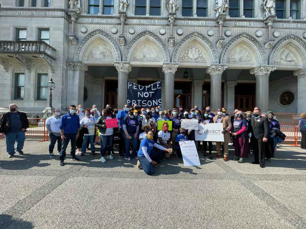 A group of Smart Justice leaders and activists with CONECT kneel and stand in front of the CT Capitol after a Clean Slate press conference. A blue "people not prisons" banner is in the background.