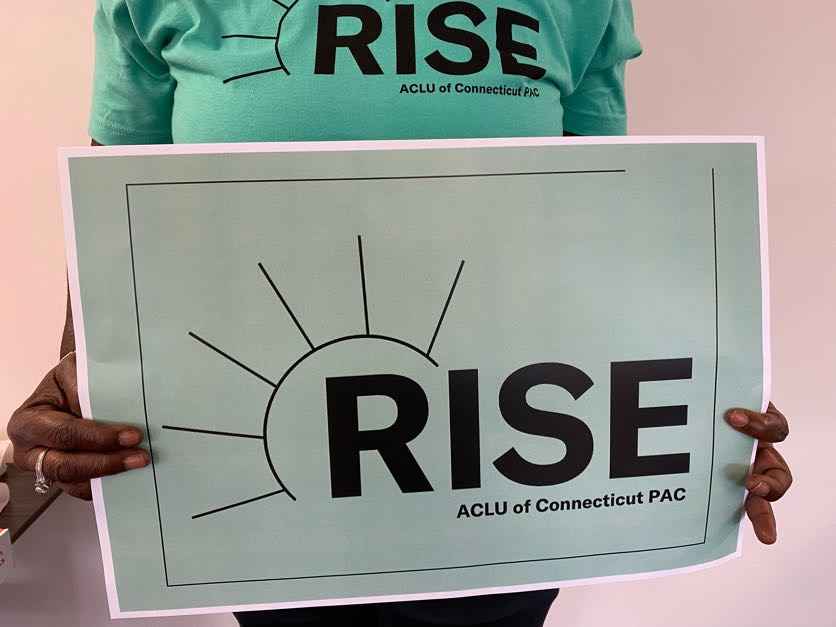 A person with long nails and a ring holds an ACLU-CT Rise PAC sign and wears an ACLU-CT Rise PAC shirt. The shirt and sign are statute of liberty green, the ACLUCT Rise PAC logos in black
