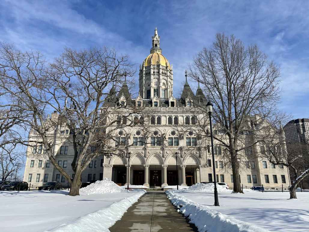 The Connecticut capitol building is centered. The capitol lawn is covered in snow, and a sidewalk leading to the building is clear of snow. The sky is blue and has light clouds.
