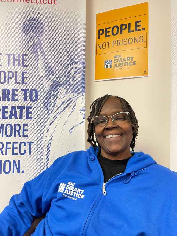 ACLU of CT Smart Justice leader Terri Ricks sits, smiling and facing the camera. She is wearing glasses and a blue people not prisons sweatshirt, in front of a yellow people not prisons sign. 